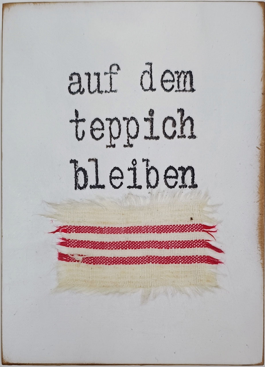 Objektkunst von Jan M. Petersen -auf dem teppich bleiben- Auf hellem Grund der Bildtitel in Schreibmaschinentypografie, darunter ein aufgeklebtes, rot-weißes Textilfragment.